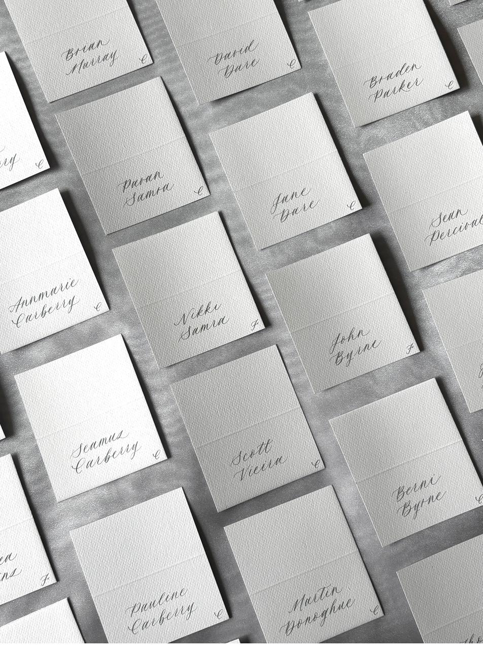 Grey and white wedding place cards laid out in a diagonal grid, each hand-lettered with a guest name