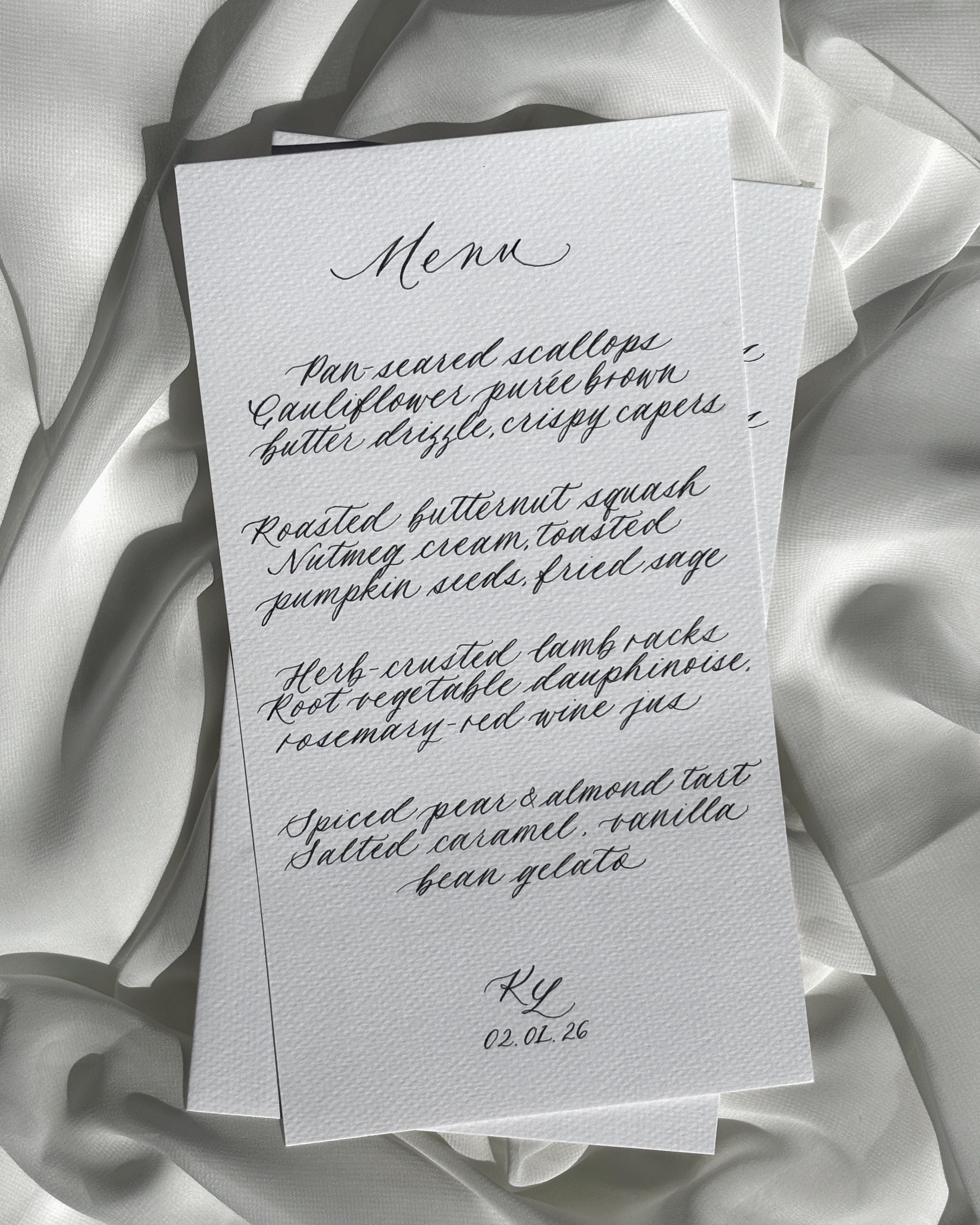 Hand-lettered dinner menu card laid on white fabric, listing a four-course meal in script