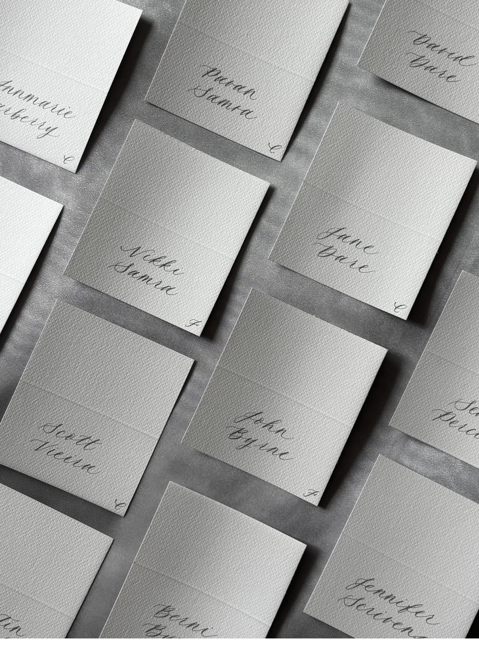 Grey and white wedding place cards laid out in a grid, each hand-lettered with a guest name